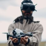 Blurred figure of man wearing tan jacket and black FPV goggles holding white and black camera drone extended toward camera in open field, showcasing compact aerial photography equipment.