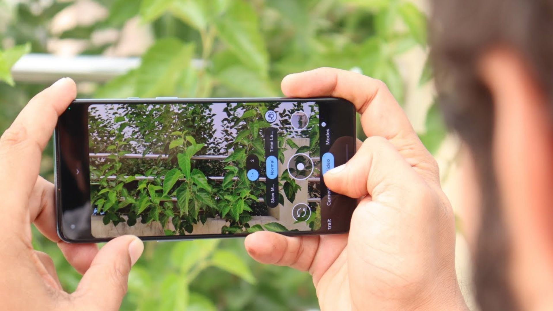 Hands holding smartphone using GCam app to photograph green plants outdoors, showing camera interface with multiple shooting modes and settings controls clearly visible.
