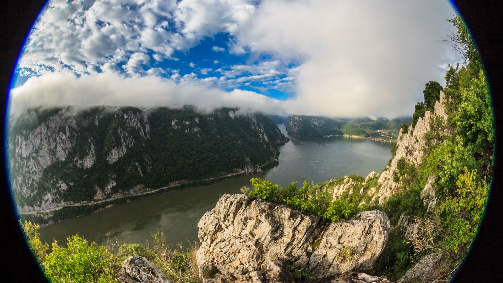Dramatic fisheye panorama of river gorge winding through steep canyon walls, low clouds rolling over mountains, rocky cliff foreground, blue sky patches, creating sweeping ultra-wide nature vista.
