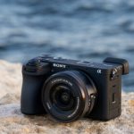 Sony α6700 with kit lens placed on sandy rock near ocean water with blurred waves in background showing outdoor environmental lifestyle photography scene.