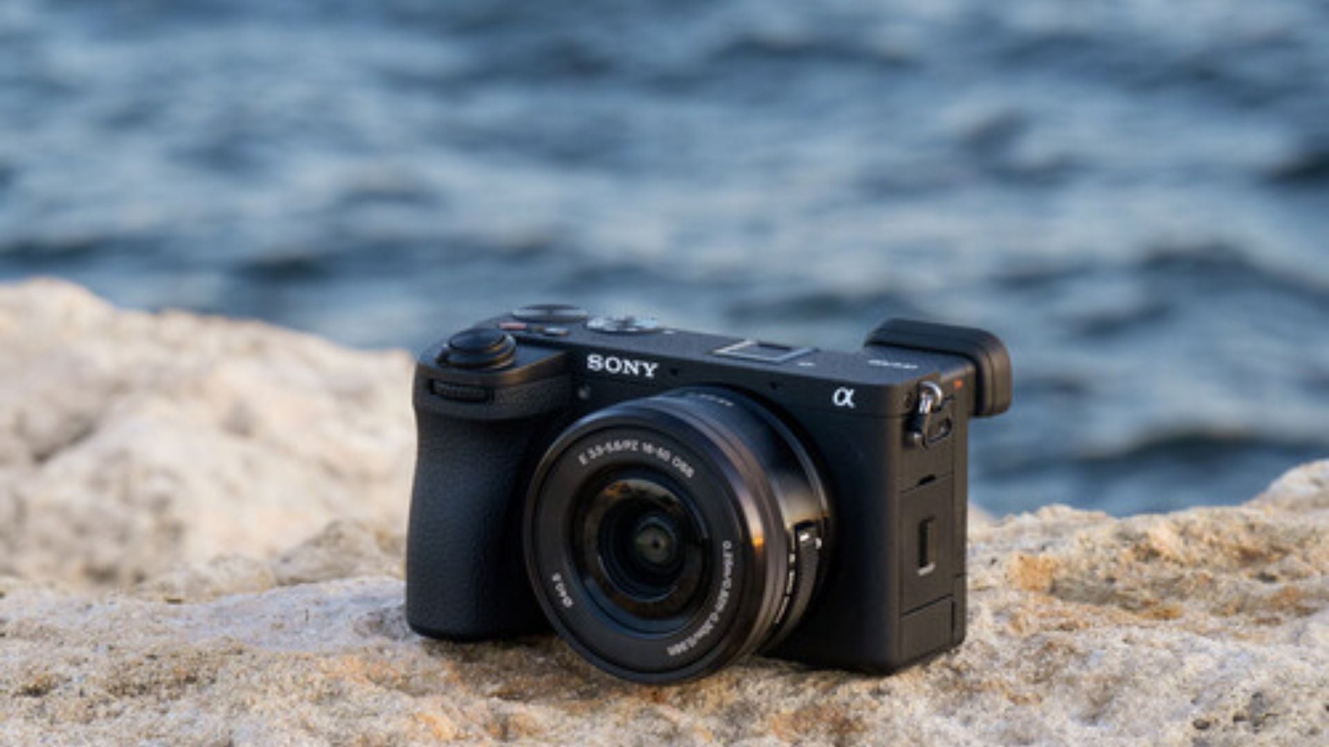 Sony α6700 with kit lens placed on sandy rock near ocean water with blurred waves in background showing outdoor environmental lifestyle photography scene.