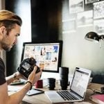 Man in brown t-shirt examining Canon DSLR camera at wooden desk, dual monitors displaying photo galleries, desk lamp, laptop, coffee mugs, and framed photography prints on wall in home office.
