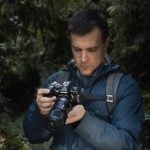 Young man in blue hooded jacket and backpack examining mirrorless camera with macro lens while standing in dense green forest, looking down at camera display reviewing nature macro photography shots.