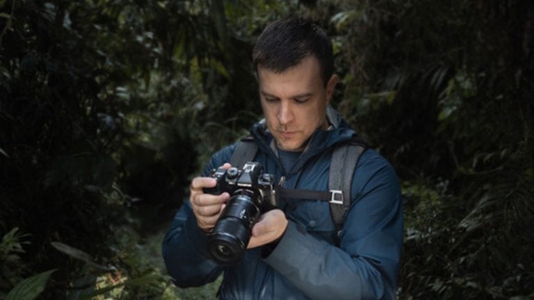 Young man in blue hooded jacket and backpack examining mirrorless camera with macro lens while standing in dense green forest, looking down at camera display reviewing nature macro photography shots.