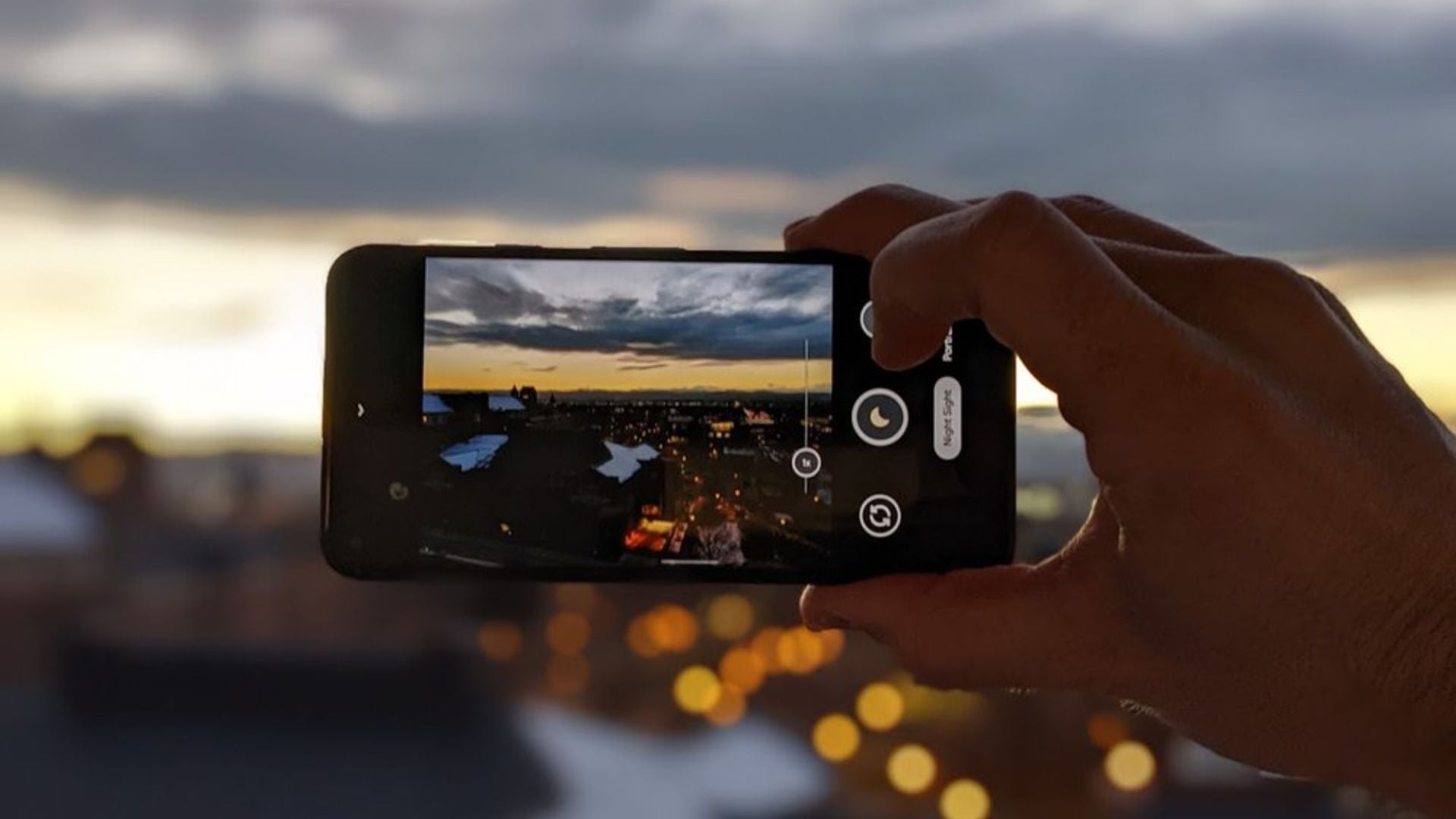Hand holding phone using GCam to capture dramatic sunset cityscape with dark clouds, golden hour lighting, and urban landscape silhouette with bokeh lights.