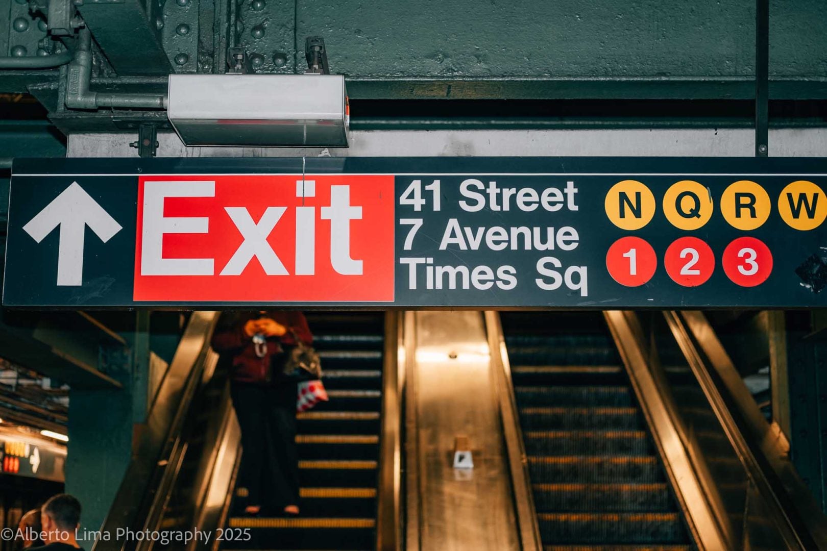A Subway sign is illuminated by a camera flash as a woman rides the escalator down.