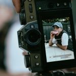 Mirrorless camera LCD displaying woman in baseball cap blowing pink bubble gum. Manual mode settings visible showing shutter speed 1/125 and ISO 200 for portrait shoot.