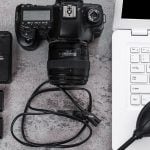 Canon DSLR camera with lens, battery charger, spare batteries, cables, and rocket blower arranged on gray surface beside white keyboard and computer for photo editing workflow.