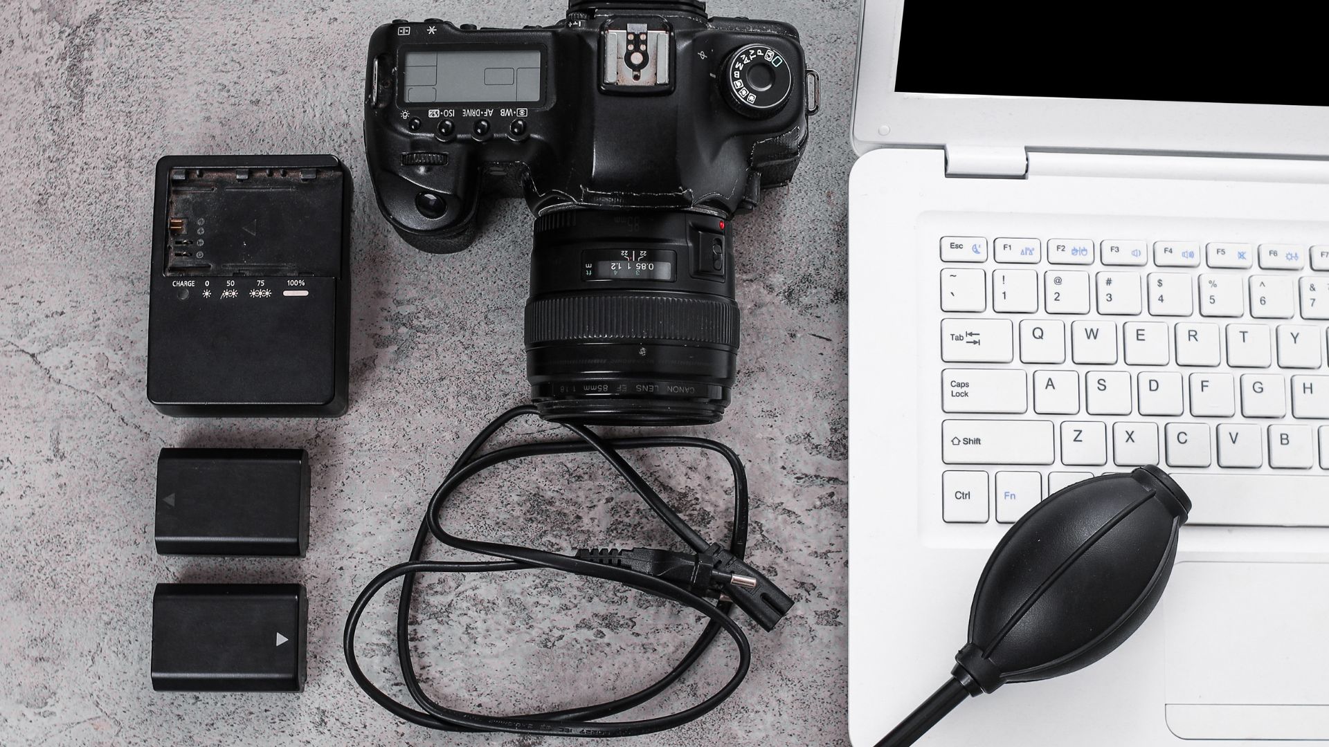 Canon DSLR camera with lens, battery charger, spare batteries, cables, and rocket blower arranged on gray surface beside white keyboard and computer for photo editing workflow.