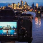 Mirrorless camera rear view showing LCD screen with illuminated city skyline during blue hour, displaying f/16 aperture, 13-second shutter speed, and ISO 50 settings