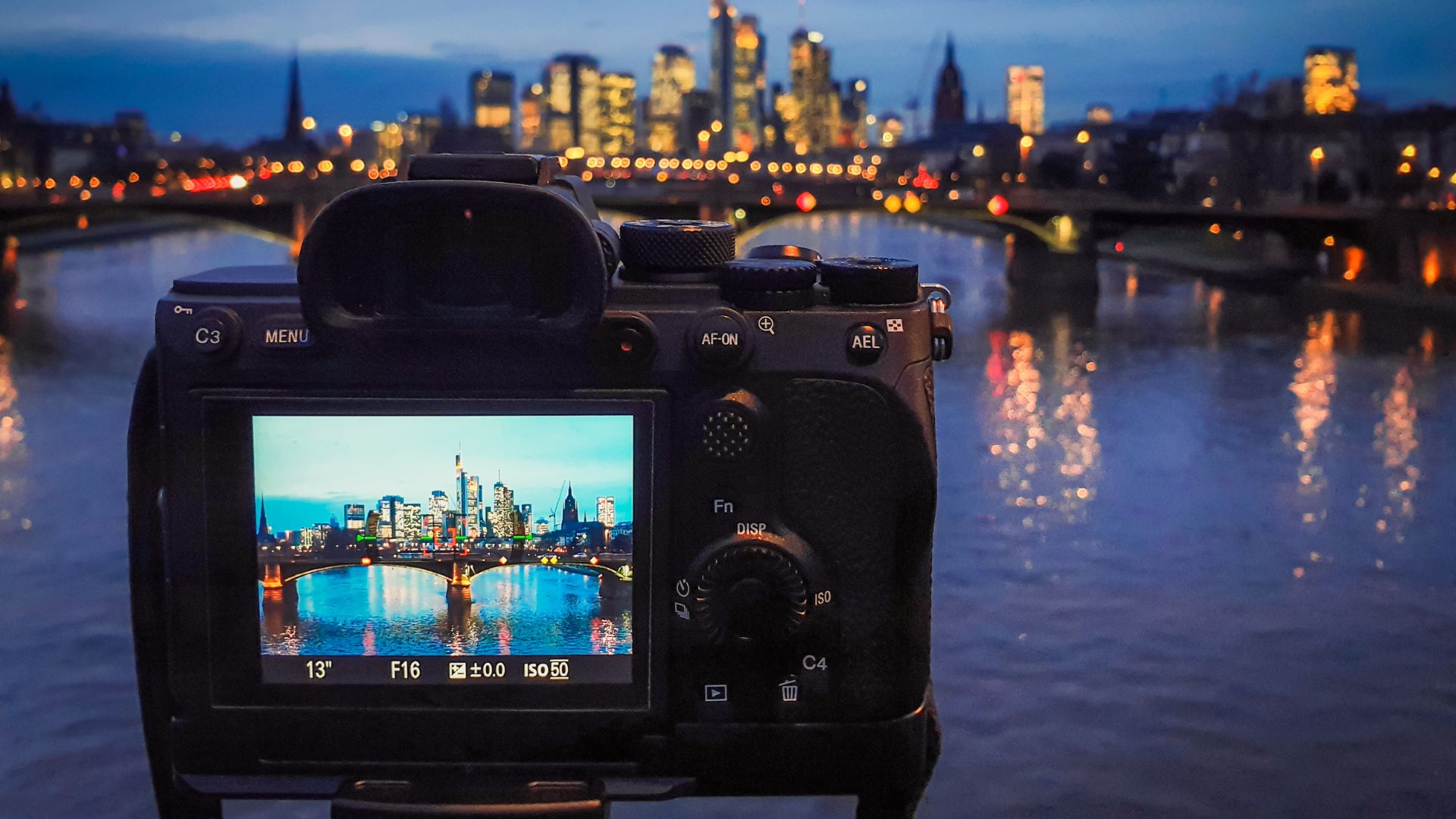 Mirrorless camera rear view showing LCD screen with illuminated city skyline during blue hour, displaying f/16 aperture, 13-second shutter speed, and ISO 50 settings