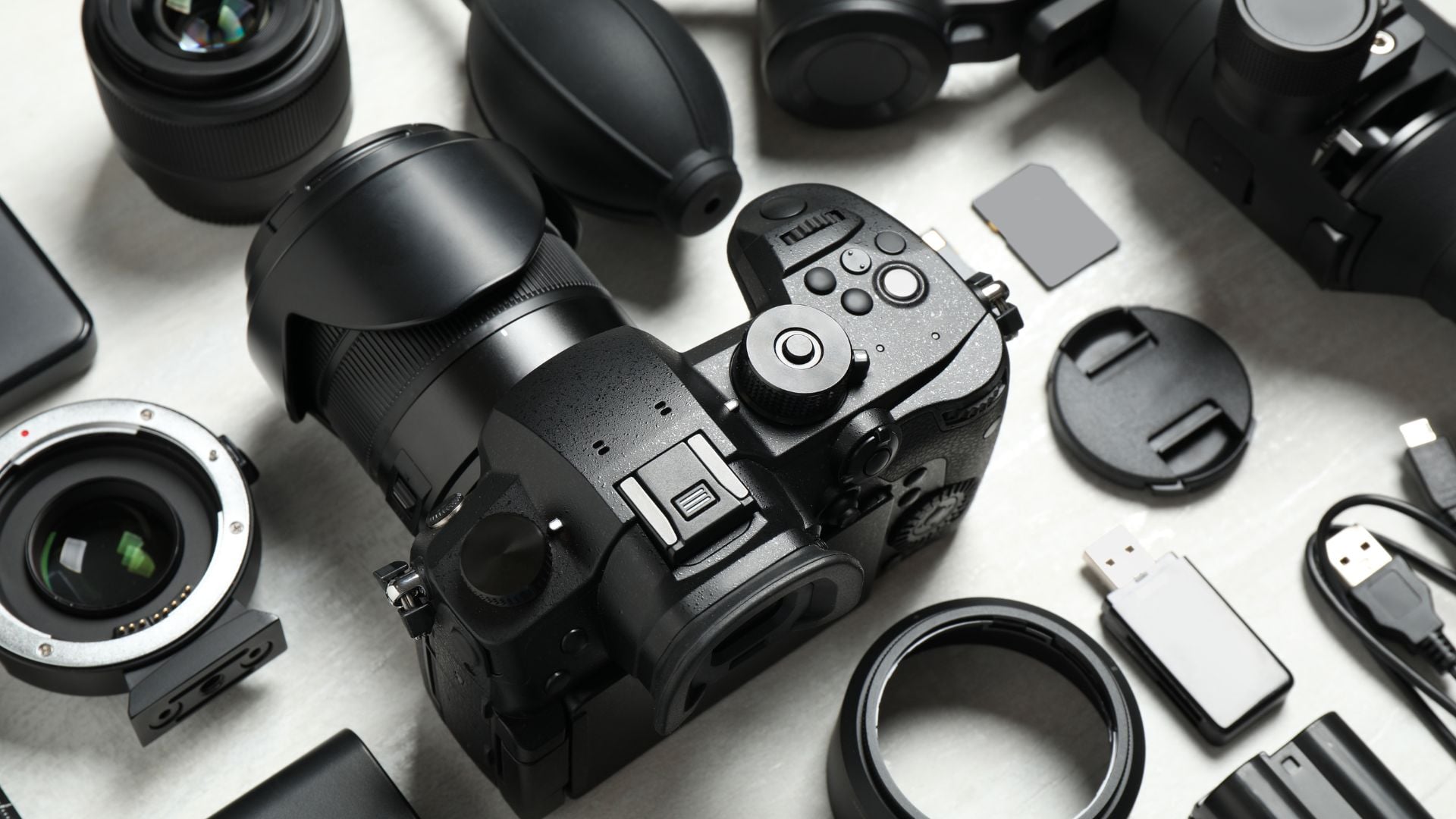 Professional photography gear including mirrorless camera body with telephoto lens, lens adapters, filters, memory cards, USB cables, and camera accessories arranged on white surface for overview.