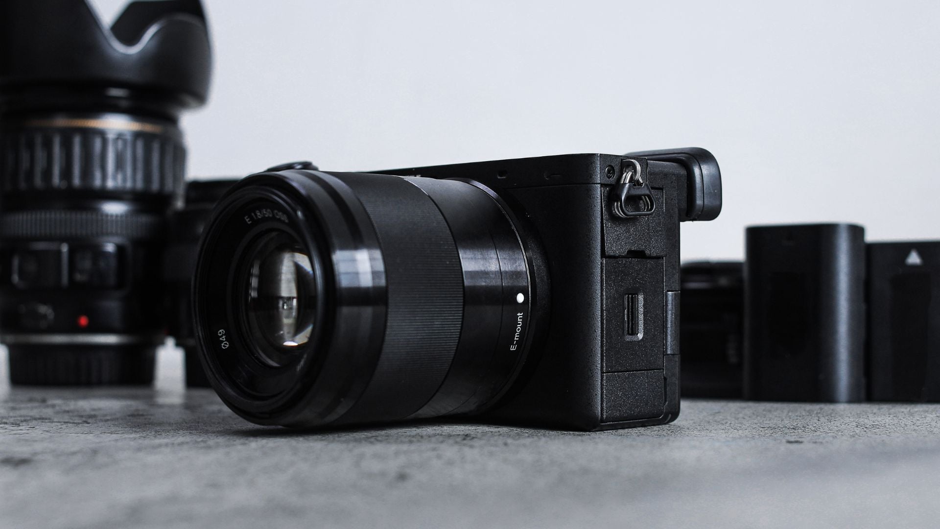 Black Sony mirrorless camera with E-mount lens shown from side angle. Compact camera body displays ports and controls with battery and additional lens visible in blurred background.