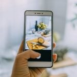 Hand holding smartphone photographing food styling setup with pineapple, cutting board, and fruit bowl demonstrating mobile food photography technique