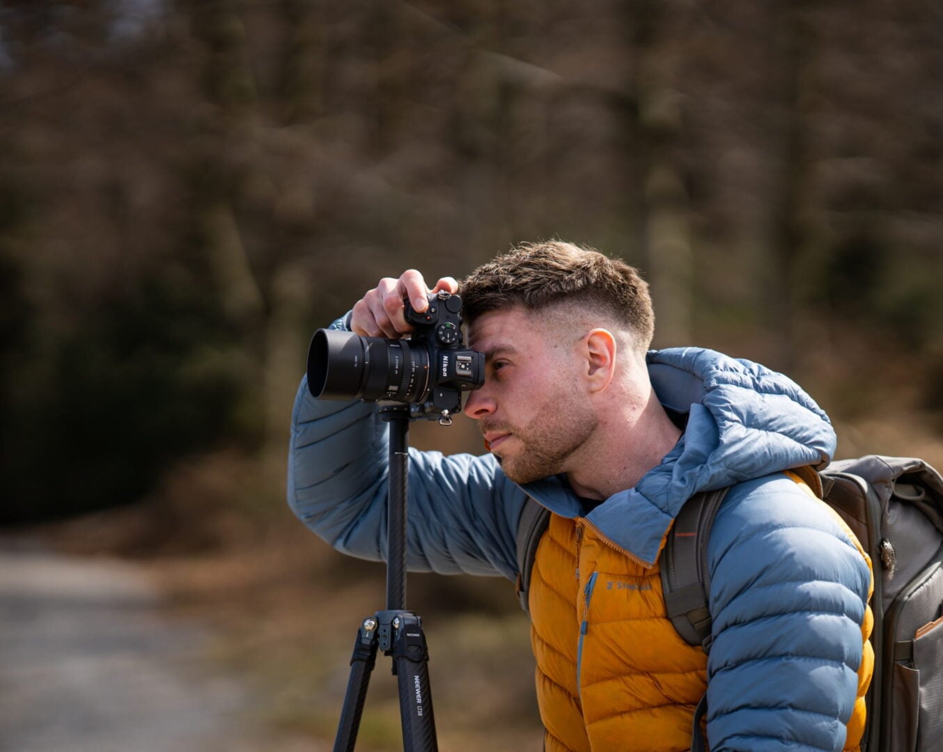 A man in hiking equipment looks through a camera viewfinder. The Viltrox AF90mm F2.2 EVO for Nikon Z cameras is mounted on the camera.