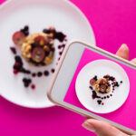 Top view close-up of female hands making flat lay food photo with phone for restaurant menu or social network. Oatmeal porridge on pink background, mockup, template, copy space, shallow depth of field